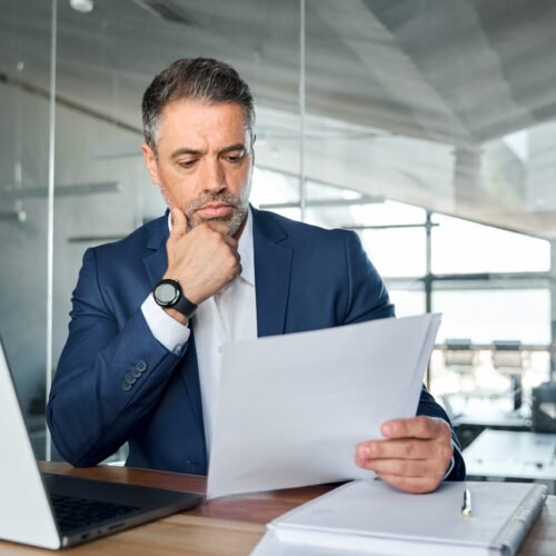 Serious busy mid aged professional business man lawyer or financial law expert wearing suit holding corporate documents reading paper contract sitting at desk in office managing risks feeling doubt.